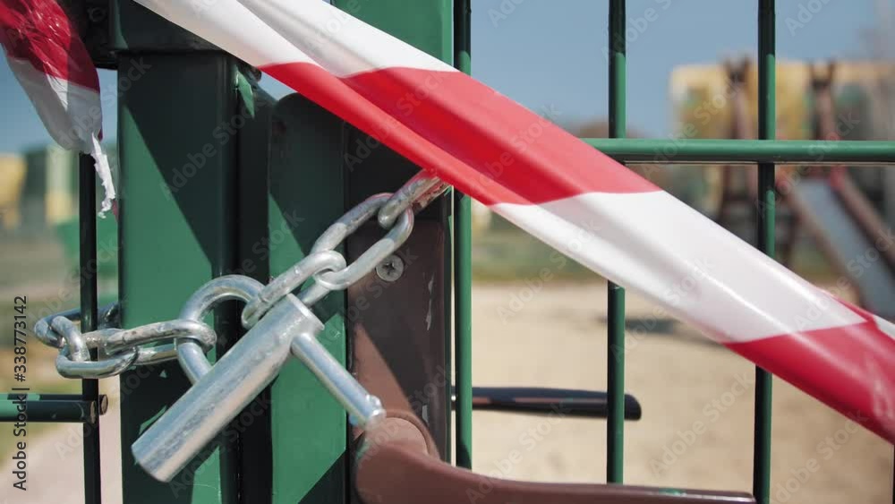 White and red tape prohibiting anyone from entering the playground. Place closed until further notice. The gate is locked with a padlock. Time coronavirus epidemic in Europe. Close Up. White and red tape prohibiting anyone from entering the playground. Place closed until further notice. The gate is locked with a padlock. Time coronavirus epidemic in Europe. Close Up.