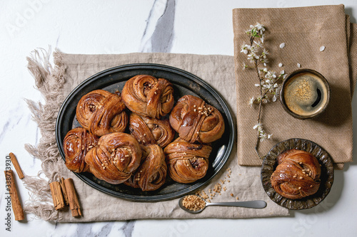 Traditional Swedish cinnamon sweet buns Kanelbulle on vintage tray, cup of coffee, cinnamon sticks, blossom branches on linen cloth over white marble background. Flat lay, space. photo