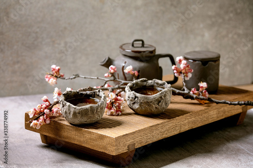 Tea drinking wabi sabi japanese style dark clay cups and teapot on wooden tea table with blooming cherry branches. Grey texture concrete background. photo