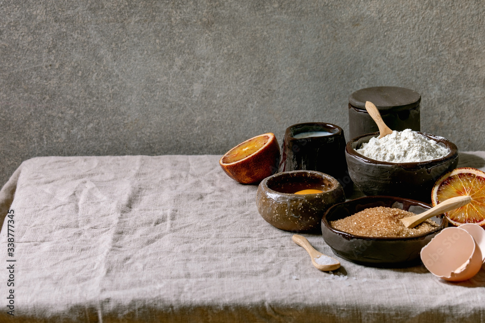 Ingredients for home baking. Flour, eggs, cane sugar, milk in different ceramic bowls, blood oranges and spices on linen table cloth. Grey texture background Ingredients for home baking. Flour, eggs, cane sugar, milk in different ceramic bowls, blood oranges and spices on linen table cloth. Grey texture background
