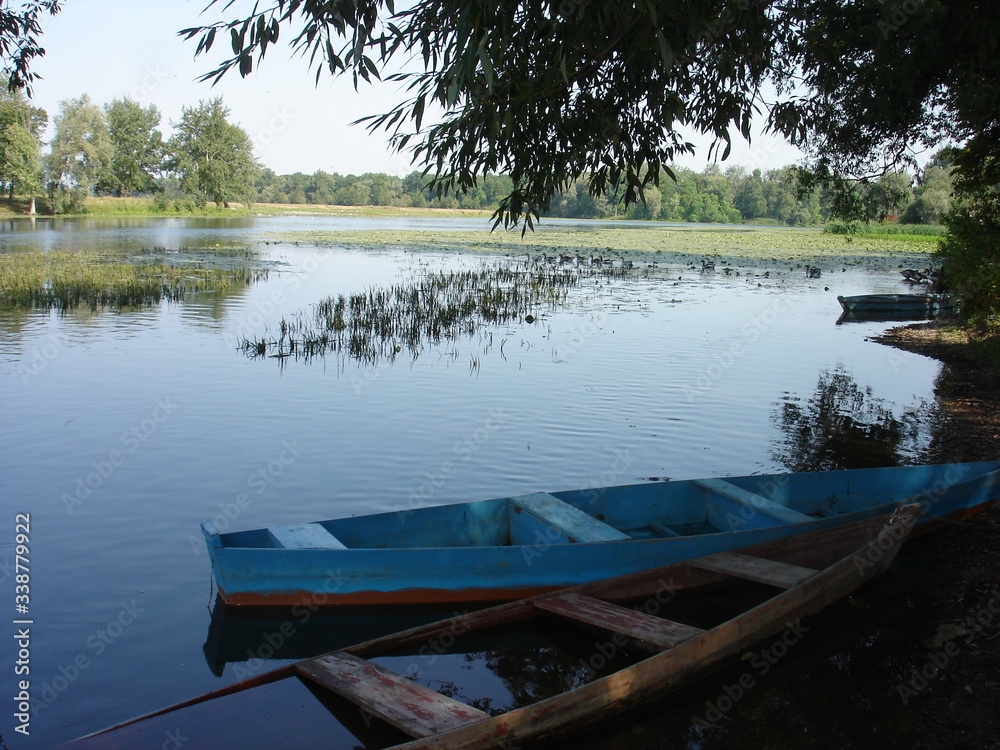 A quiet, calm river overgrown with water vegetation. Early morning. Two old wooden boats are moored on the shore.