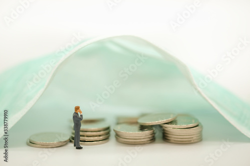 Global Healthcare, Coronavirus, Covid-19 Protection Concept. Businessman miniature figure looking for pile silver coins under sugical mask.