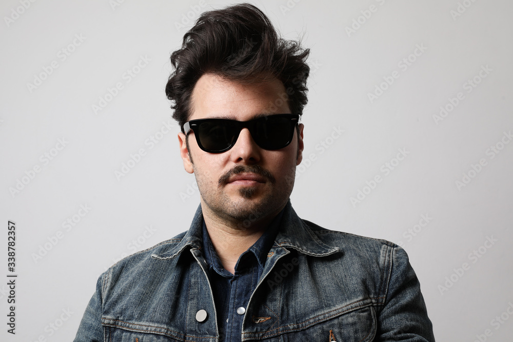 Obraz premium Close-up portrait of serious stylish hipster looks with confident expression, wears sunglasses and denim jacket.