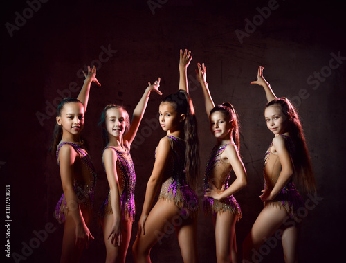 Team of young smiling girls gymnasts in purple shiny sport body posing over grey background