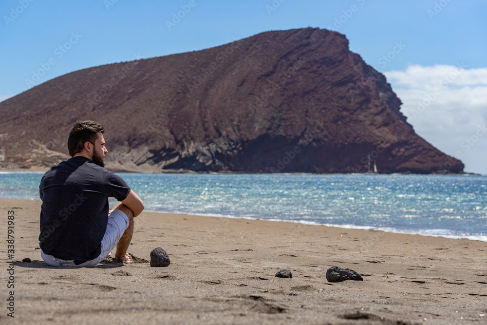 A young handsome man sitting on the sand in a beach while looking at the sea, in Tenerife, Canary Islands, Spain