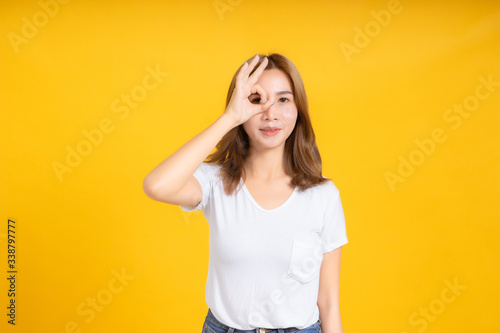 Portrait young happy asian woman ok gesture hand your eye symbol for advertising promotion sale discount in white t-shirt, Yellow background isolated studio shot and copy space.