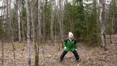 a blonde girl in a green jumpsuit runs and dances in the spring forest.a lot of trees and leaves, energetic movements, Christmas trees are nearby, a pink hat and gloves