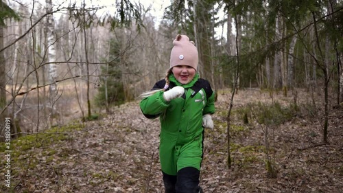 a blonde girl in a green jumpsuit runs and dances in the spring forest.a lot of trees and leaves, energetic movements, Christmas trees are nearby, a pink hat and gloves.she runs around the trees