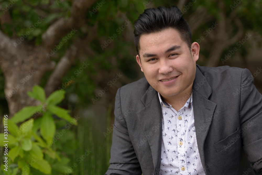 Happy young overweight Asian businessman smiling at the park Happy young overweight Asian businessman smiling at the park