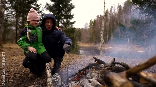 Camping: little girl is sitting on her father's lap next to the fire.They are in jackets, on the street autumn or spring, trees river and forest surround them, the girl has green outfit.They talking