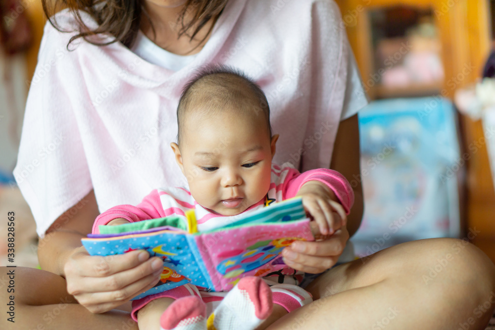 Obraz premium 6-month-old Asian girl sat on the mother's lap and reading a children's story book.