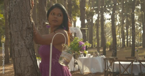 An east indian model standing in a pine forest with decorated wedding tables in the background at this outdoor wedding event photo