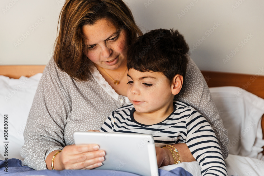una mujer madre y su hijo con pluridiscapacidad (minusvalía, discapacidad) juegan con el ordenador portátil o tablet en la cama. van en pijama, y están alegres y sonrientes.