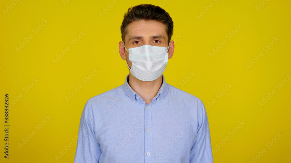 Man in protective medical mask looks at the camera. The guy in the blue shirt on a yellow background. Concept against the virus: Coronavirus, flu Man in protective medical mask looks at the camera. The guy in the blue shirt on a yellow background. Concept against the virus: Coronavirus, flu