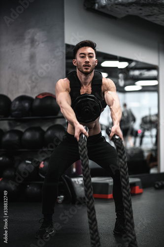 Man during workout in the gym Concept: power, strength, healthy lifestyle, sport. Powerful attractive muscular Man CrossFit trainer do battle workout with ropes at the gym. Young man exercising using