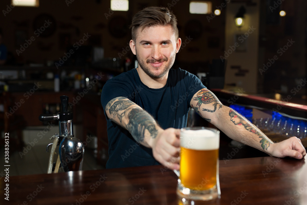 Handsome young male bartender in stretching out glass with beer and smiling while standing at the bar counter