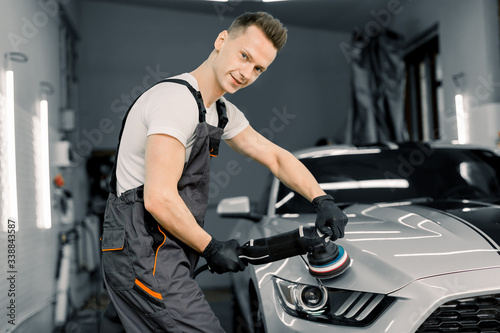 Young handsome smiling Caucasian man, worker of auto detailing service, holds a polisher in the hand and polishes the car, looking and posing at camera. Car detailing and polishing concept