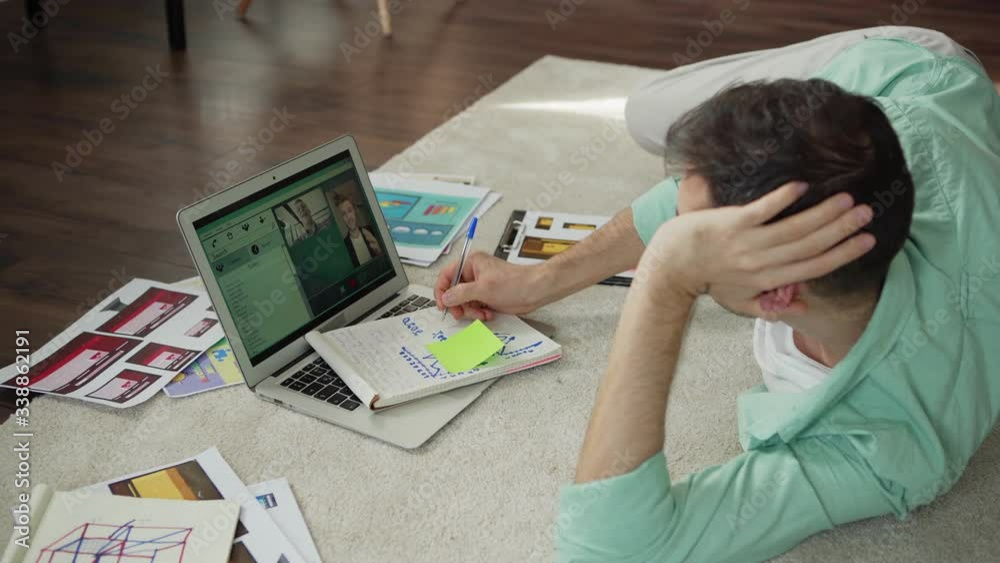 Panning shot of young man greeting two male colleagues and discussing business during video call on laptop computer lying on floor at home surrounded by papers Panning shot of young man greeting two male colleagues and discussing business during video call on laptop computer lying on floor at home surrounded by papers