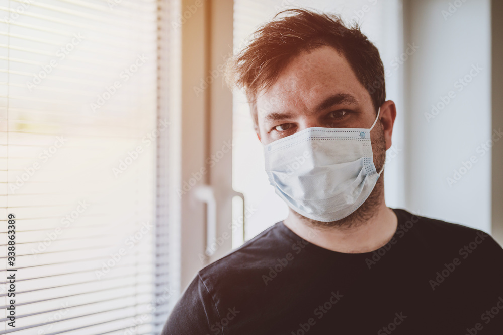 Man in quarantine during coronavirus COVID-19. Man with protective mask stands near window. Novel coronavirus 2019 COVID-19 theme. Quarantine during coronavirus pandemic Man in quarantine during coronavirus COVID-19. Man with protective mask stands near window. Novel coronavirus 2019 COVID-19 theme. Quarantine during coronavirus pandemic