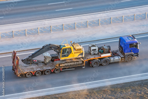 Truck with a long trailer platform for transporting heavy machinery, loaded crawler excavator with bucket. Highway transportation.