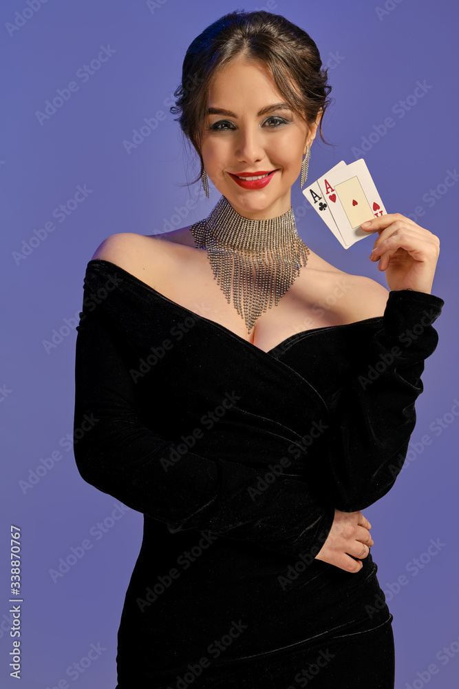 Brunette lady with bare shoulders, in black dress and shiny jewelry. Smiling, showing two aces, posing on purple background. Poker, casino. Close-up Brunette lady with bare shoulders, in black dress and shiny jewelry. Smiling, showing two aces, posing on purple background. Poker, casino. Close-up