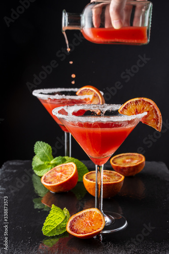 Top view of two cocktail glasses with unfocused bottle serving drops of blood orange martini, half orange and mint, on black background, vertical