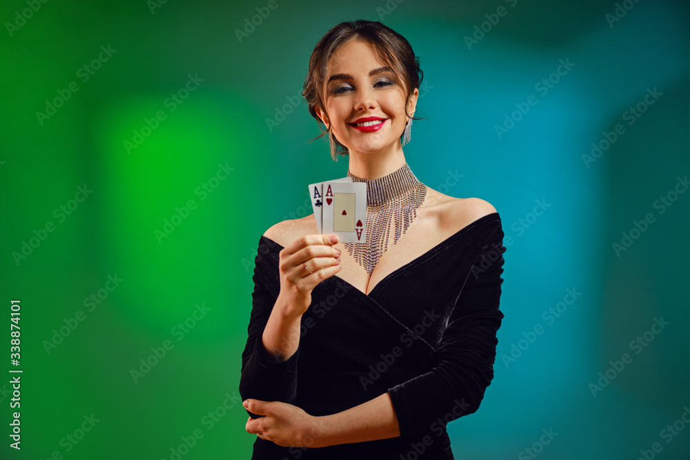 Brunette girl in black dress, shiny necklace and earrings. Smiling, showing two playing cards, posing on colorful background. Poker, casino. Close-up Brunette girl in black dress, shiny necklace and earrings. Smiling, showing two playing cards, posing on colorful background. Poker, casino. Close-up