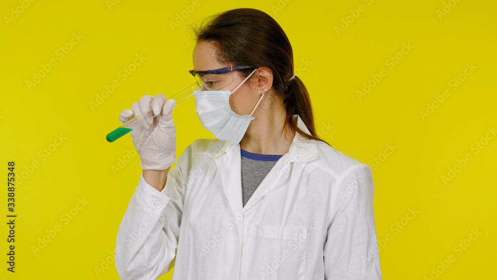 Doctor in a medical mask, goggles and latex gloves looks at the tests in the tube. A young girl in a white coat on a yellow background holds hand a tube with green liquid