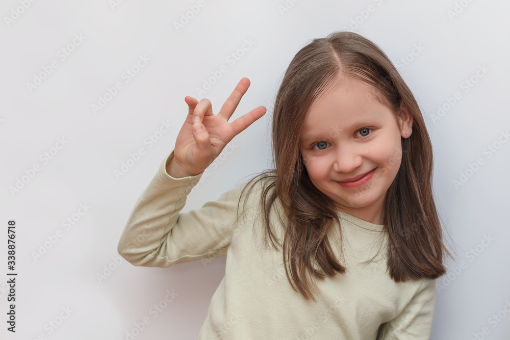 Little girl in a light sweater on a white gray background looks at the camera and shows a victory sign with her hand Little girl in a light sweater on a white gray background looks at the camera and shows a victory sign with her hand