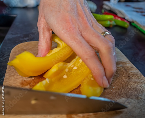 Close up of human hand slicing a yellow bell pepper (Capsicum annuum) with a sharp kitchen knife on a wooden cutting board with selective focus