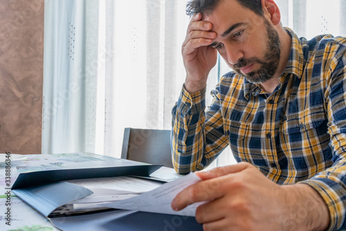 Bearded man dressed in plaid shirt looking at bills worried in his living room at home