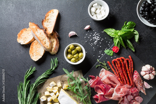 Tapas concept. Prosciutto, sausage chorizo, olives, baguette, arugula, basil and brie cheese on a black slate board. Antipasti concept.