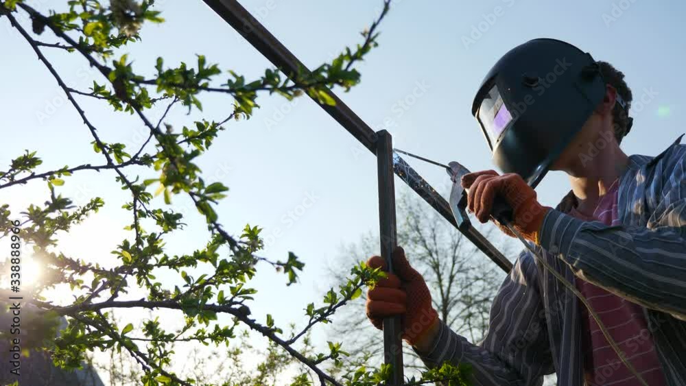 Young Man Welding Metal Construction in Backyard. Worker in Protective Helmet. Garden Near Cottage House in Countryside. 4K 2x Slow motion 0.5 x 60 FPS Young Man Welding Metal Construction in Backyard. Worker in Protective Helmet. Garden Near Cottage House in Countryside. 4K 2x Slow motion 0.5 x 60 FPS