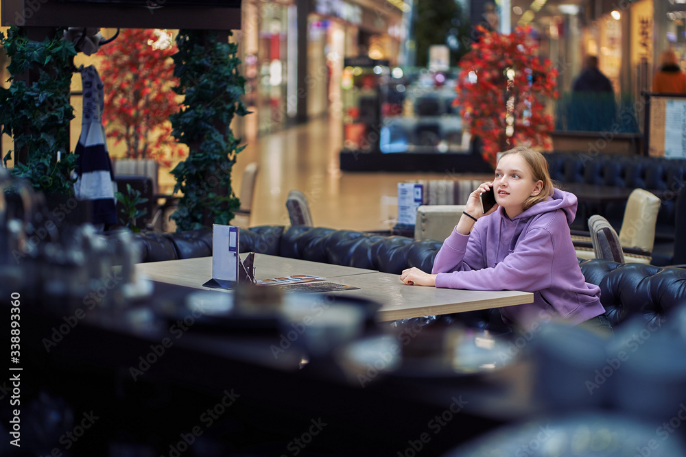 A nice girl is sitting at a table in a cafe waiting for a menu and talking on the phone.