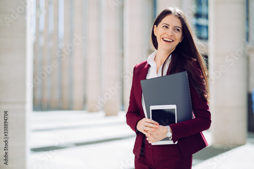 Portrait of business woman dressed in maroon suit, holds tablet and folder, smiling widely outdoor. Business people concept.
