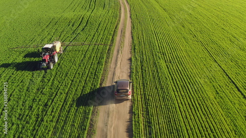 Aerial view of a farmer driving a car on a dirt road between agricultural fields. A tractor spraying fertilizer passes along the field. Drone chases a car