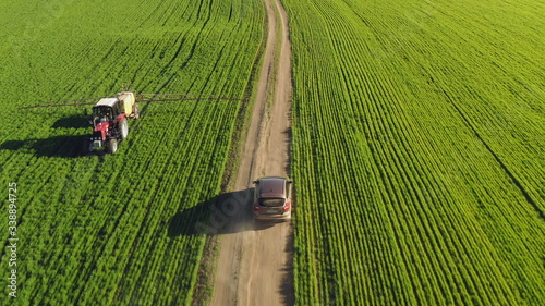 Aerial view of a farmer driving a car on a dirt road between agricultural fields. A tractor spraying fertilizer passes along the field. Drone chases a car