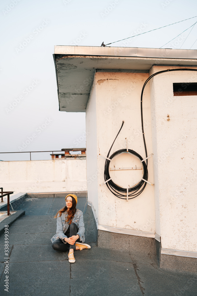 portrait pretty white European woman is sitting on the roof of a multi-storey building in a blue denim jacket in gray pants,a yellow stylish hat and yellow sneakers. lifestyle concept