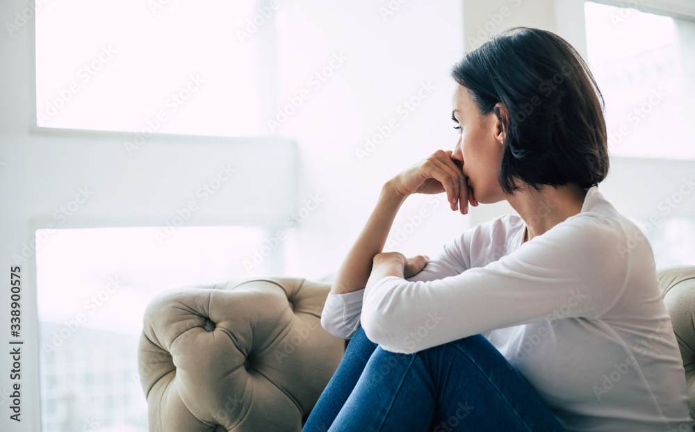 Trying to cope. Close-up photo of an adult woman who turned away from the camera, looking through the window with sadness. Trying to cope. Close-up photo of an adult woman who turned away from the camera, looking through the window with sadness.