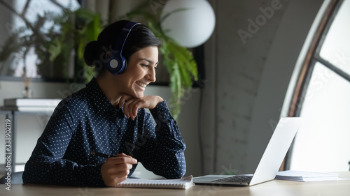 Smiling Indian female employee in wireless earphones talk on video call, have web conference with colleagues, happy ethnic woman worker in headset watch webinar on modern laptop at home photo