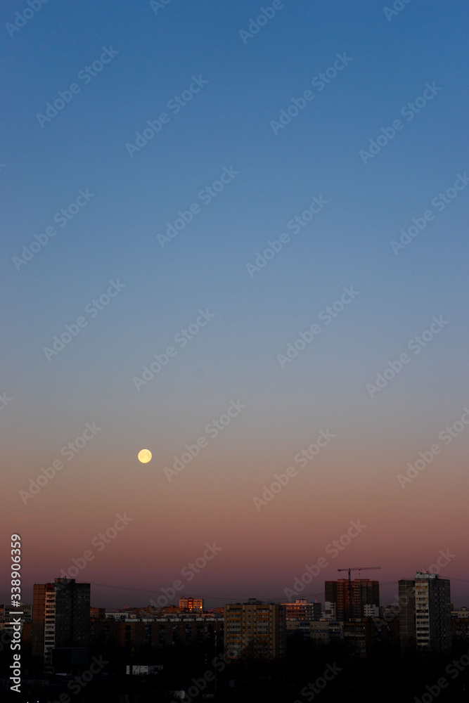 Fototapeta premium Full moon on clear sunrise sky and buildings under it. Blue and pink colours.