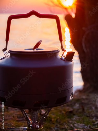 Boiling water in a touristic kettle outdoors next to the water at sunset. (ID: 338909108)
