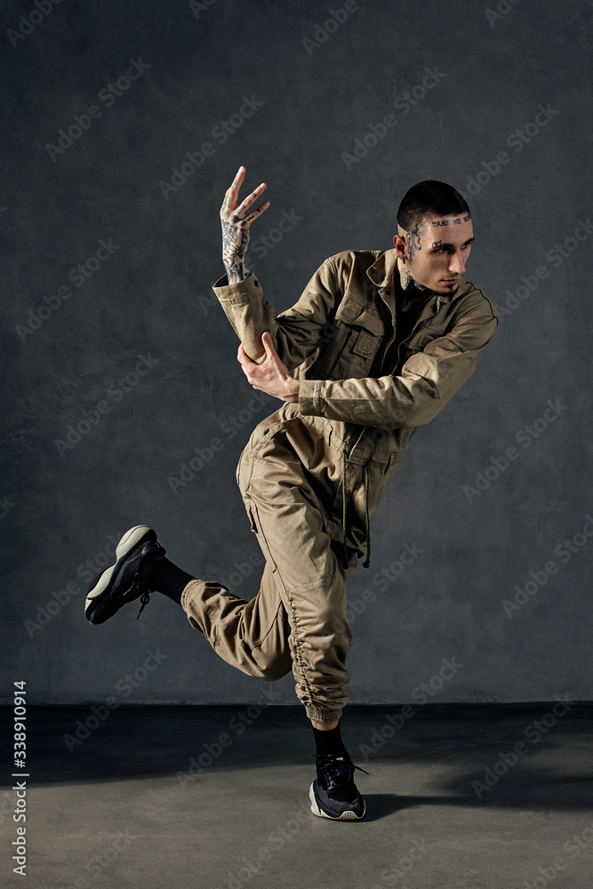 Modern man with tattooed body and face, earrings, beard. Dressed in khaki overalls and black sneakers. Dancing on gray background. Dancehall, hip-hop