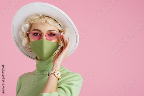 Woman wearing trendy fashion outfit during quarantine of coronavirus outbreak. Model dressed protective stylish handmade face mask, pink sunglasses, white hat, wrist watch, green mint color turtleneck