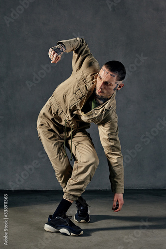 Flexible guy with tattooed body and face, earrings, beard. Dressed in khaki overalls, black sneakers. Dancing on gray background. Dancehall, hip-hop