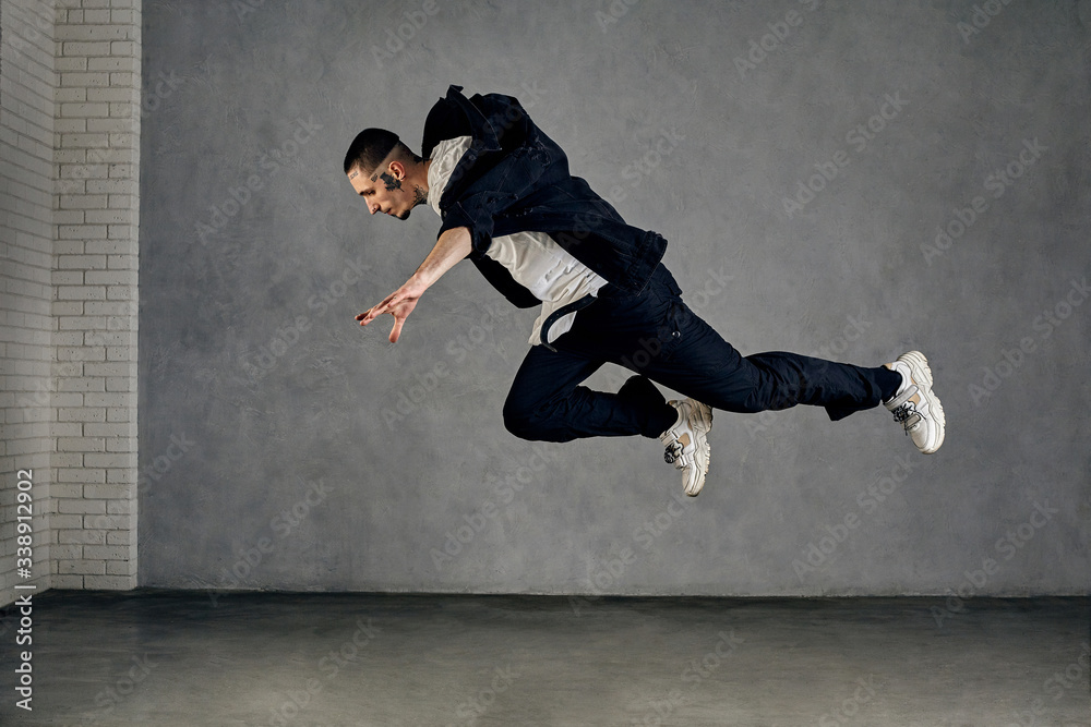 Guy with tattooed body, beard. Dressed in white t-shirt and sneakers, black shirt, pants. Performing tricks on gray background. Dancehall, hip-hop