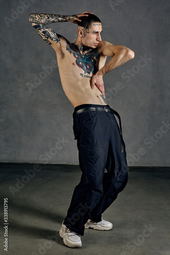 Flexible tattooed performer with naked torso, beard. Dressed in black pants and white sneakers. Dancing against gray background. Dancehall, hip-hop © nazarovsergey