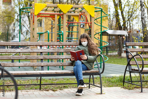 A masked teenage girl is reading a book in the playground.