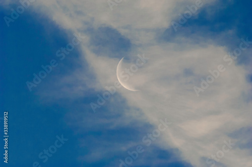 A slim sliver of the silvery moon set on an expanse of a partly cloudy blue sky.