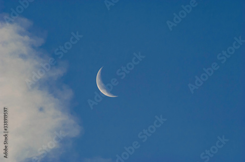 A slim sliver of the silvery moon set on an expanse of a partly cloudy blue sky.